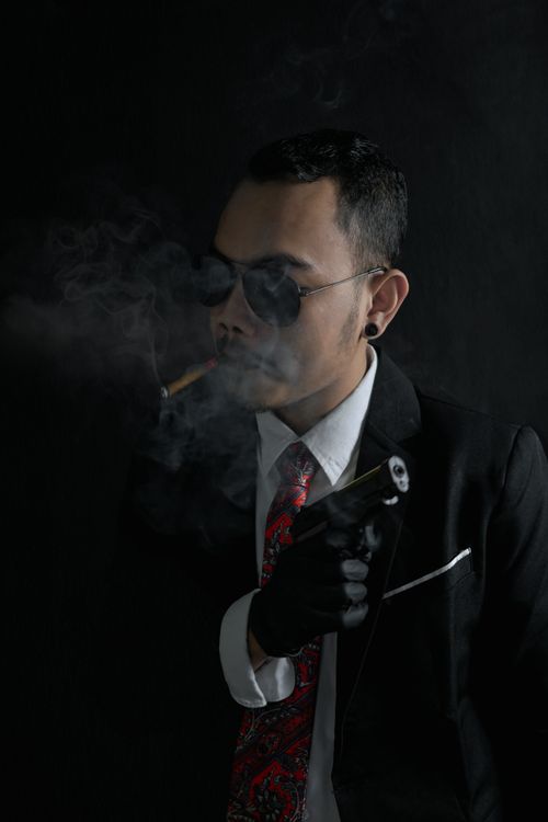 Free A man in a suit and sunglasses holding a gun while smoking in a dark studio setting. Stock Photo