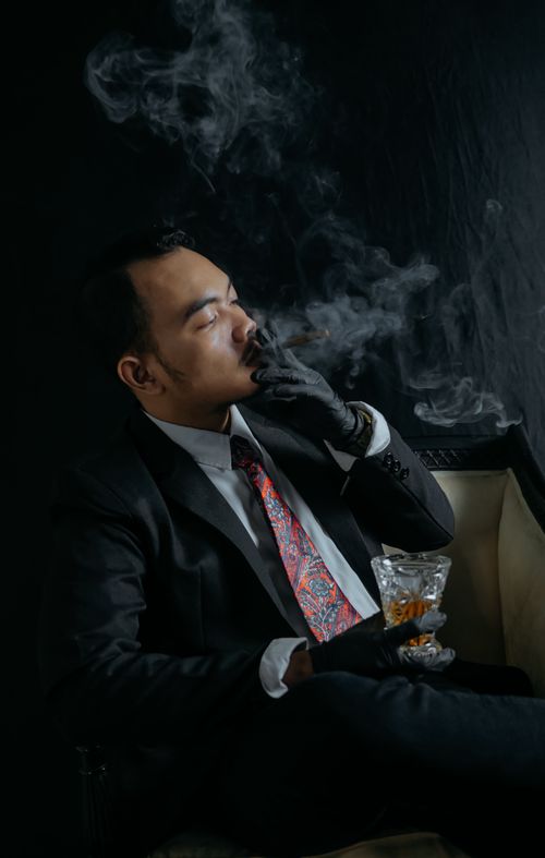 Free A man in a black suit smoking a cigar while holding a glass of whiskey in a dark setting. Stock Photo