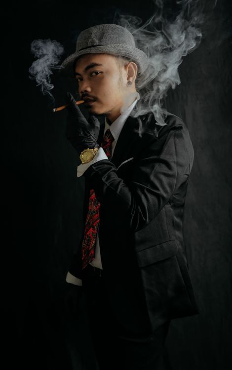Free Asian man in black suit, smoking with a gray hat and red necktie, showcasing fashion in a studio setting. Stock Photo