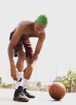 A topless young man with green hair ties his shoe on a sunny outdoor basketball court.