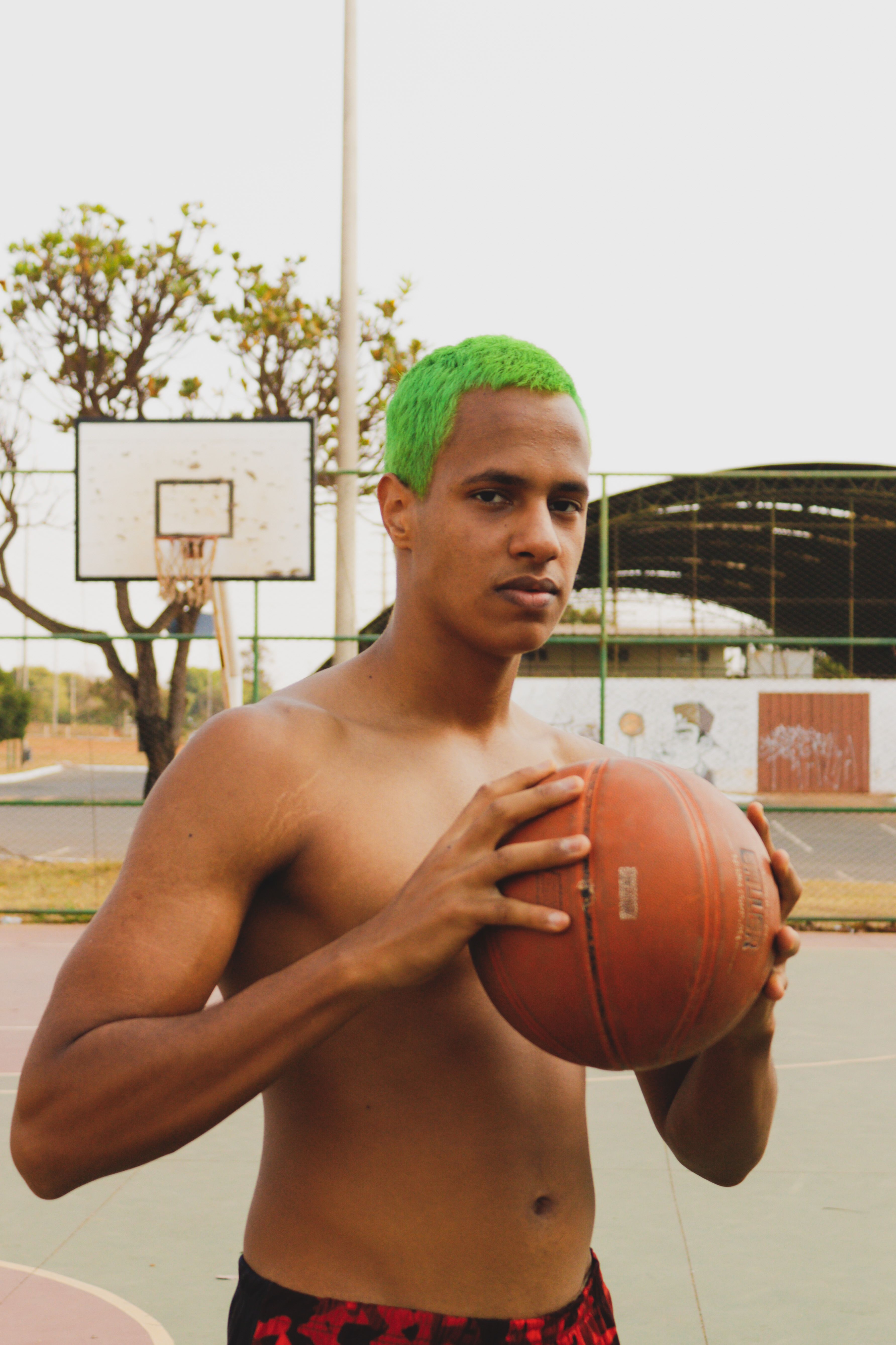 A shirtless man with green hair holds a basketball on an outdoor court.