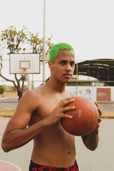 A shirtless man with green hair holds a basketball on an outdoor court.