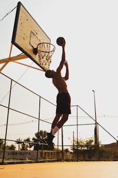 Shirtless athlete dunking basketball on outdoor court in sunlight.