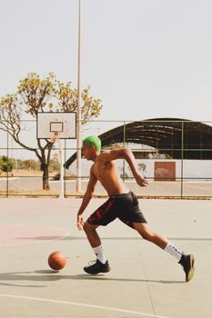Shirtless man with green hair dribbling a basketball on an outdoor court under clear skies.