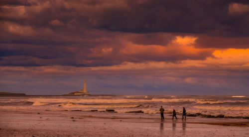 Free A scenic beach with a lighthouse and people walking under a dramatic twilight sky. Stock Photo