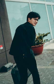 Man with eyeglasses walking on a sidewalk, holding a suitcase in a city setting.