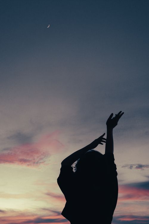 Free Silhouette of a woman with raised arms against a colorful twilight sky and crescent moon. Stock Photo