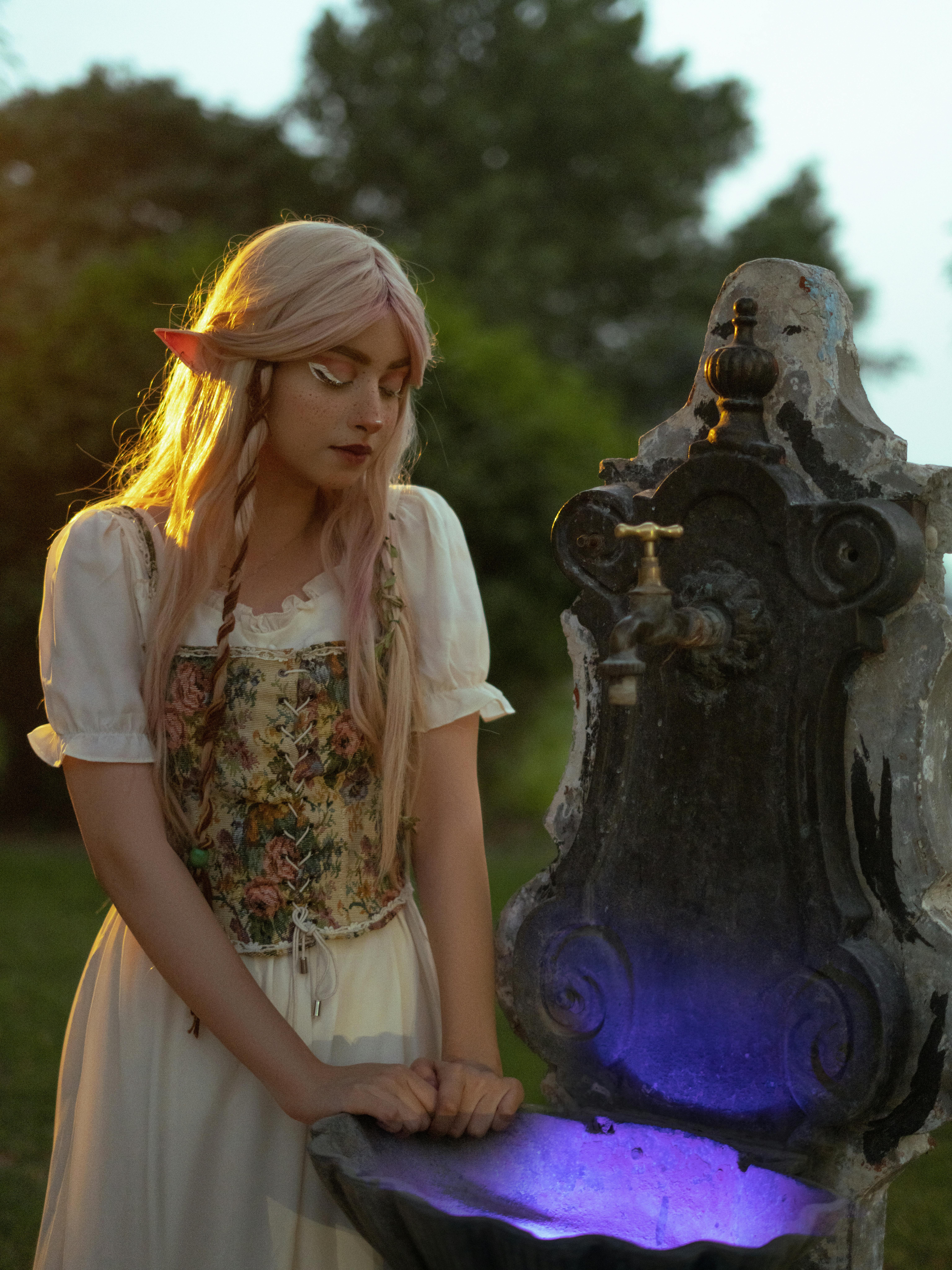 A woman in elf cosplay stands by a glowing well, evoking a magical fantasy scene.