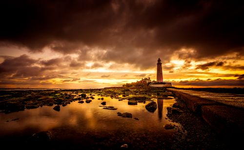 Free A stunning lighthouse stands against a vibrant sunset with dramatic clouds and rocky shoreline reflections. Stock Photo
