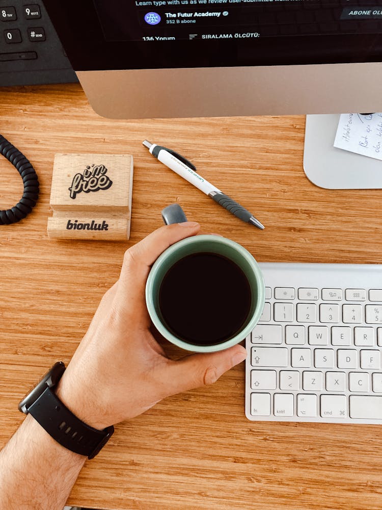 Close-up Of A Man Holding A Cup Of Coffee Next To A Computer On A Desk 