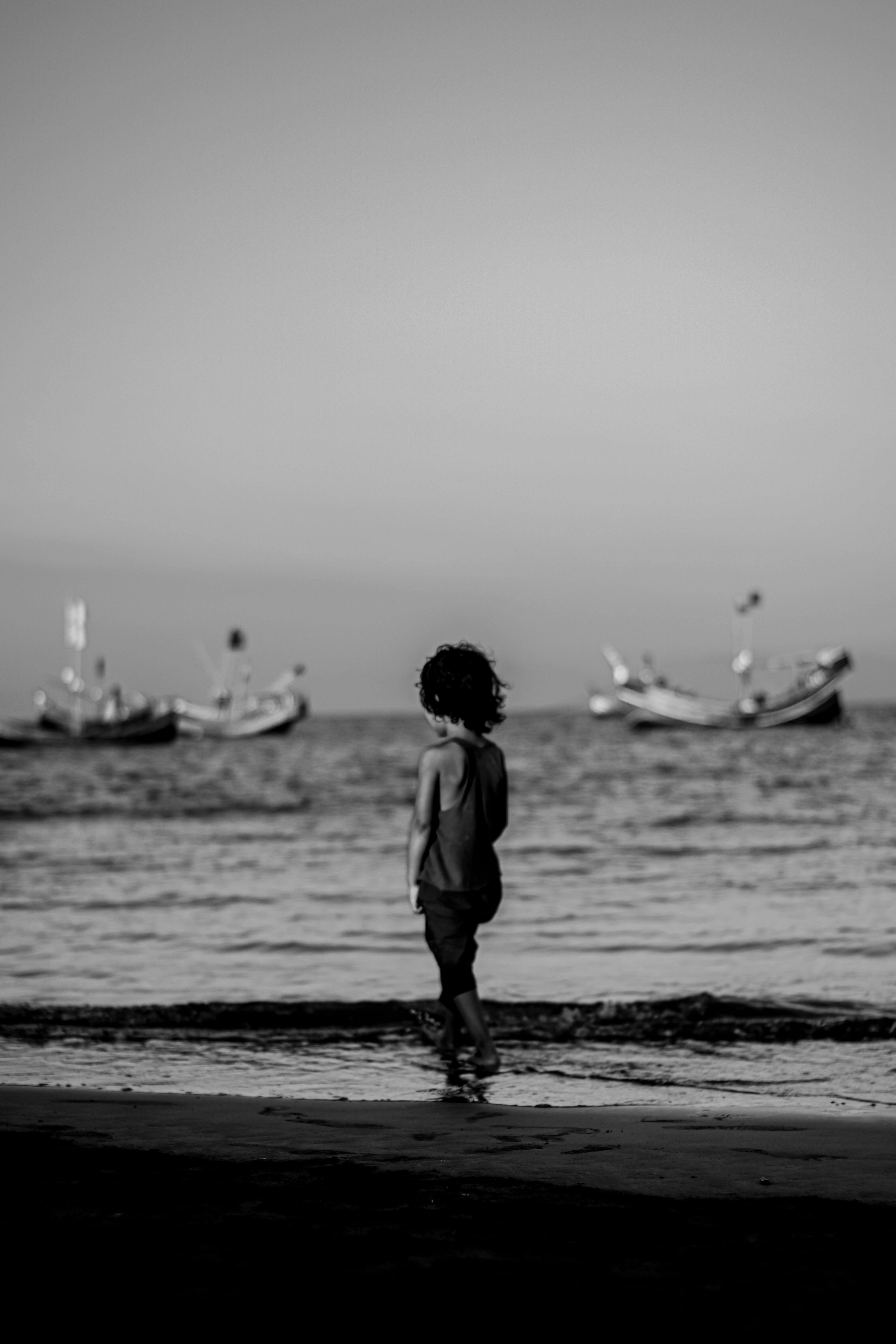 A young child standing by the sea, boats in the distance, captured in monochrome.