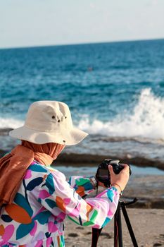 Woman in colorful attire photographs ocean waves with a DSLR camera by the shore.