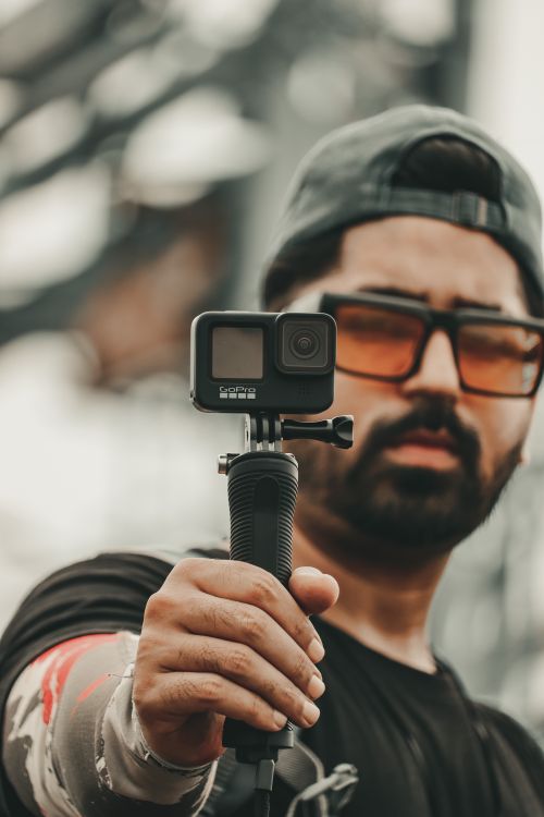 Free Man wearing cap and sunglasses holding a GoPro camera, capturing outdoor footage. Stock Photo