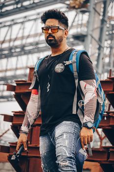Man with backpack and sunglasses in an industrial environment, wearing casual attire.