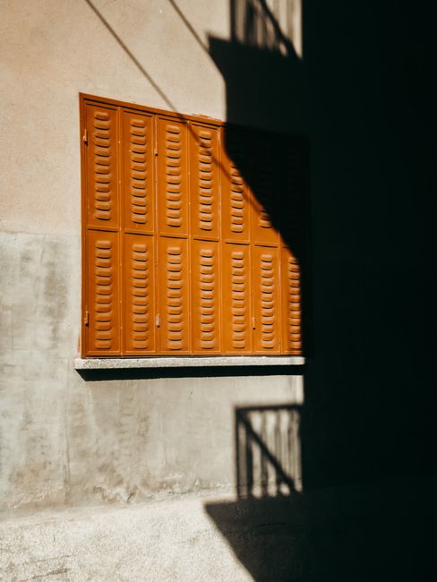 Orange shuttered window with bold shadows on a wall.