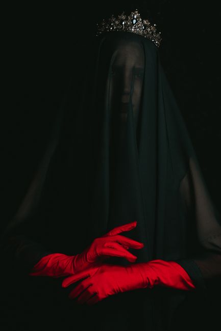 A mysterious woman in a dark veil wearing red gloves and a tiara, set against a black background.