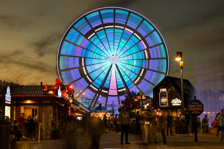 People Walking On Street Near Blue And White Ferris Wheel During Night Time