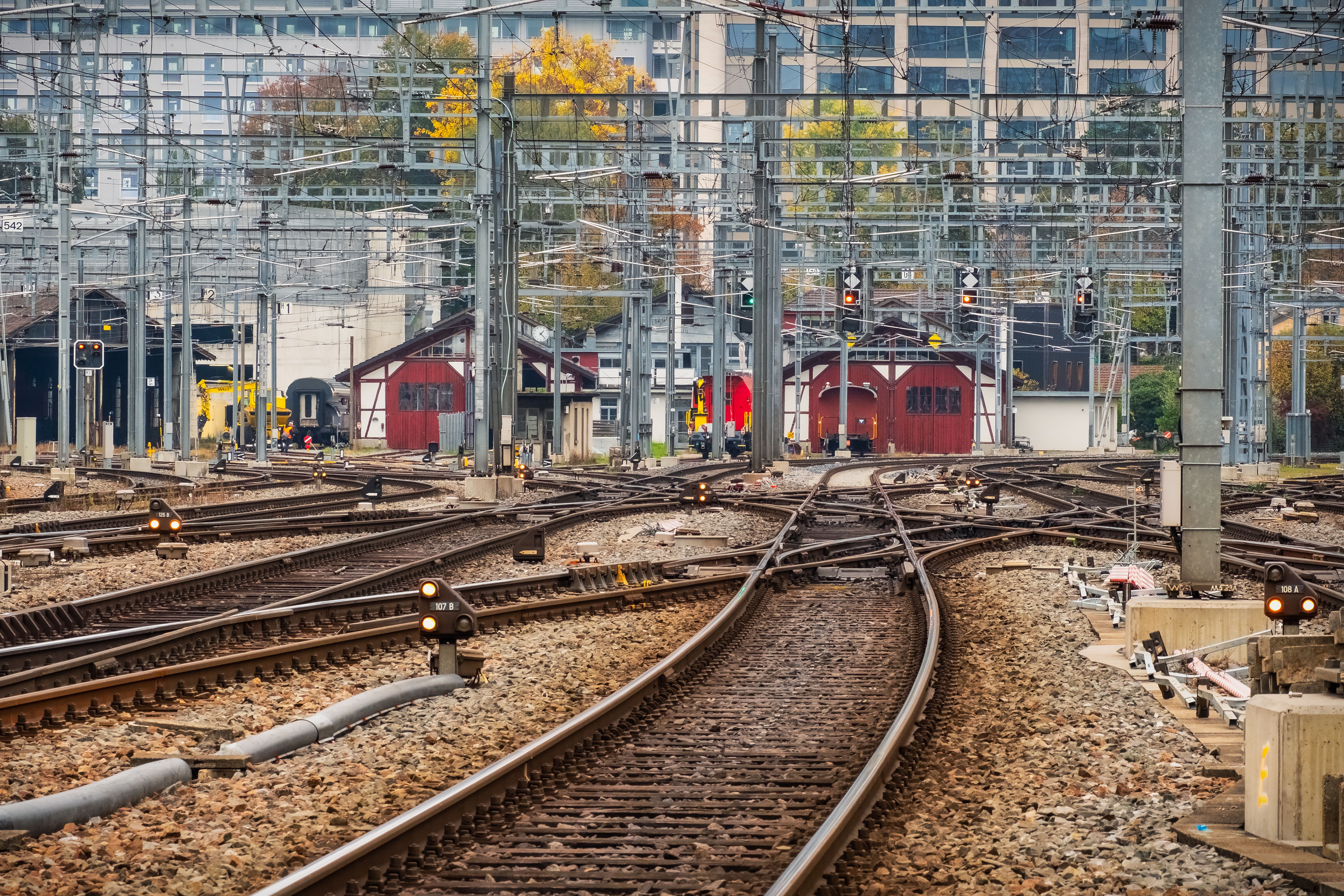 Multiple railway tracks and overhead power lines stretch through a busy urban rail yard.