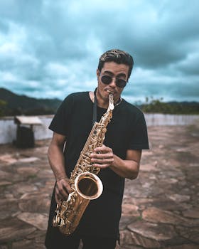Stylish male saxophonist performing outdoors with sunglasses and a cloudy sky backdrop.