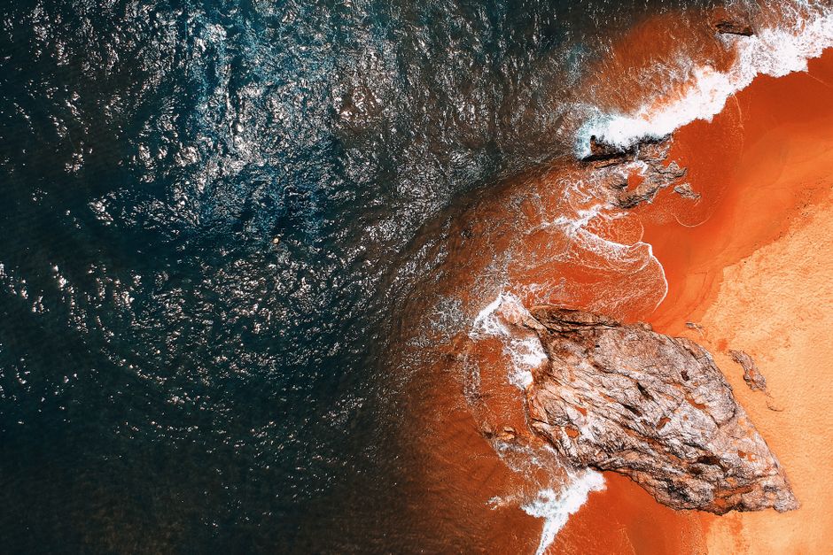 A stunning aerial shot of a vibrant seashore with waves crashing against rocky formations and orange sands.