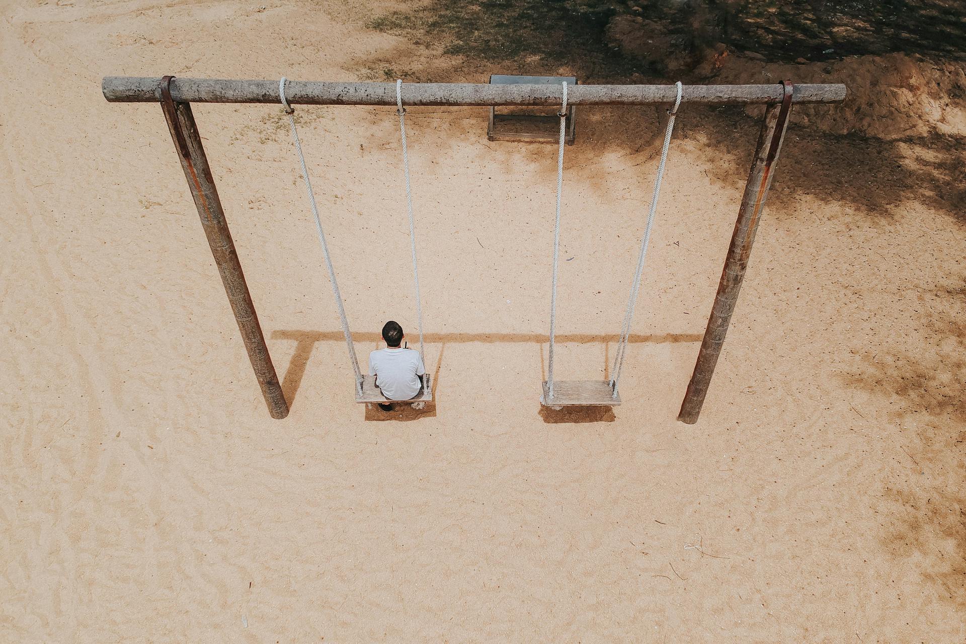 Aerial shot of a man sitting alone on a swing in a sandy playground, viewed from above.