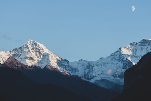 Capture the breathtaking snow-covered Swiss Alps with a serene half-moon sky.