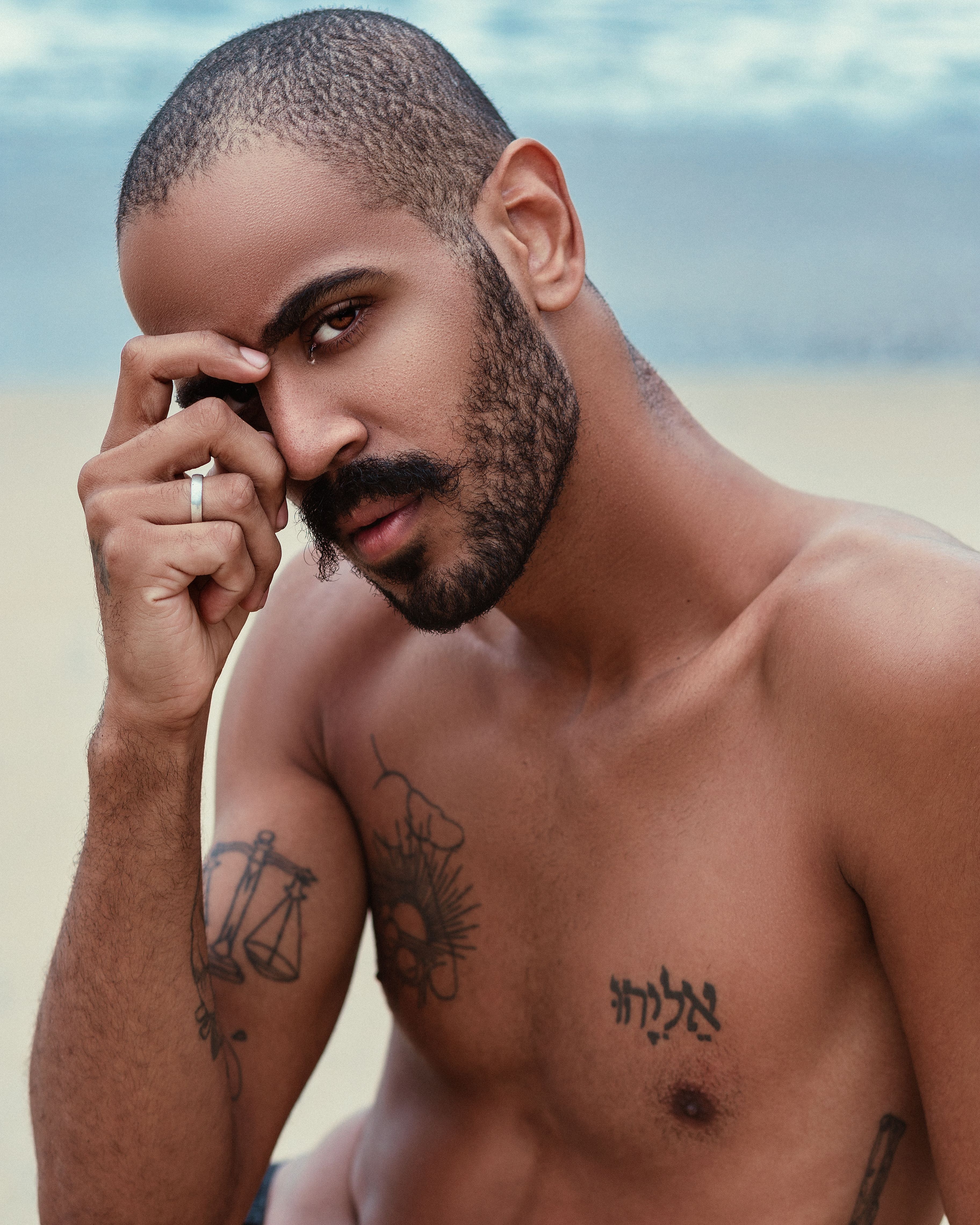 Close-up shot of a shirtless bearded man with tattoos by the sea, featuring a contemplative pose.