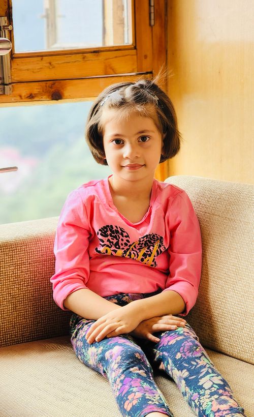Free A cute young girl sitting on a sofa indoors, wearing a bright outfit with a cheerful smile. Stock Photo