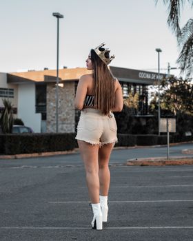 Backview of a woman wearing a crown, heels, and casual attire in a parking lot at twilight.