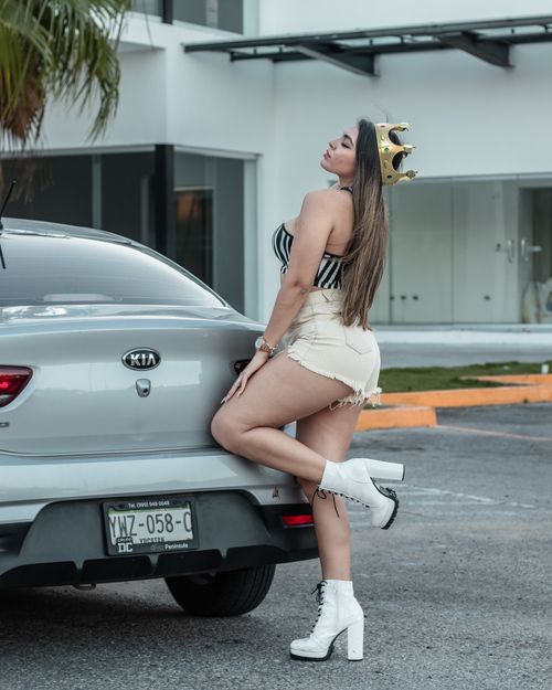 Free Confident woman wearing a crown poses with a car in a modern outdoor setting. Stock Photo