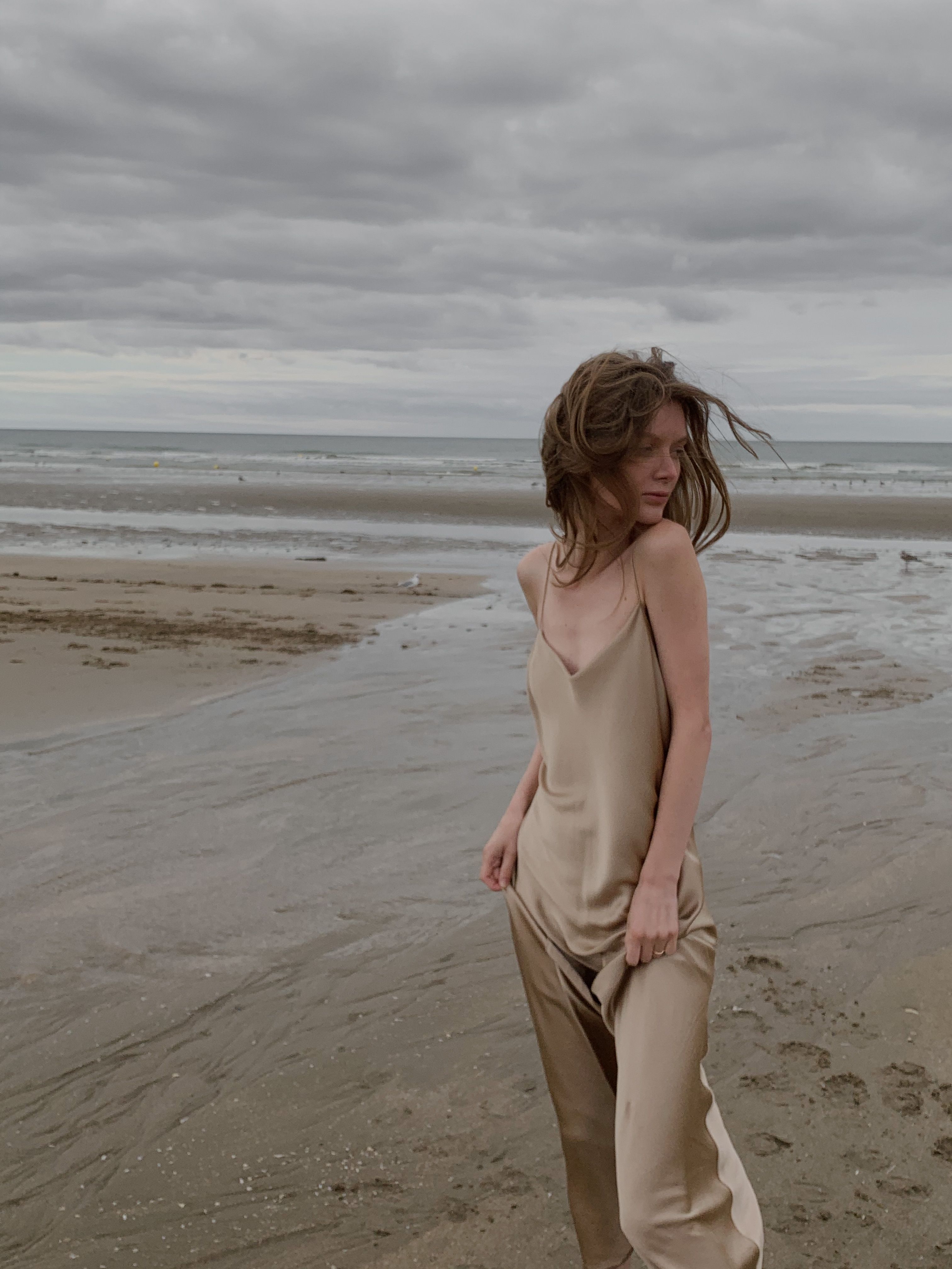 A woman in a flowing beige dress stands on a cloudy beach, facing sideways with windblown hair.