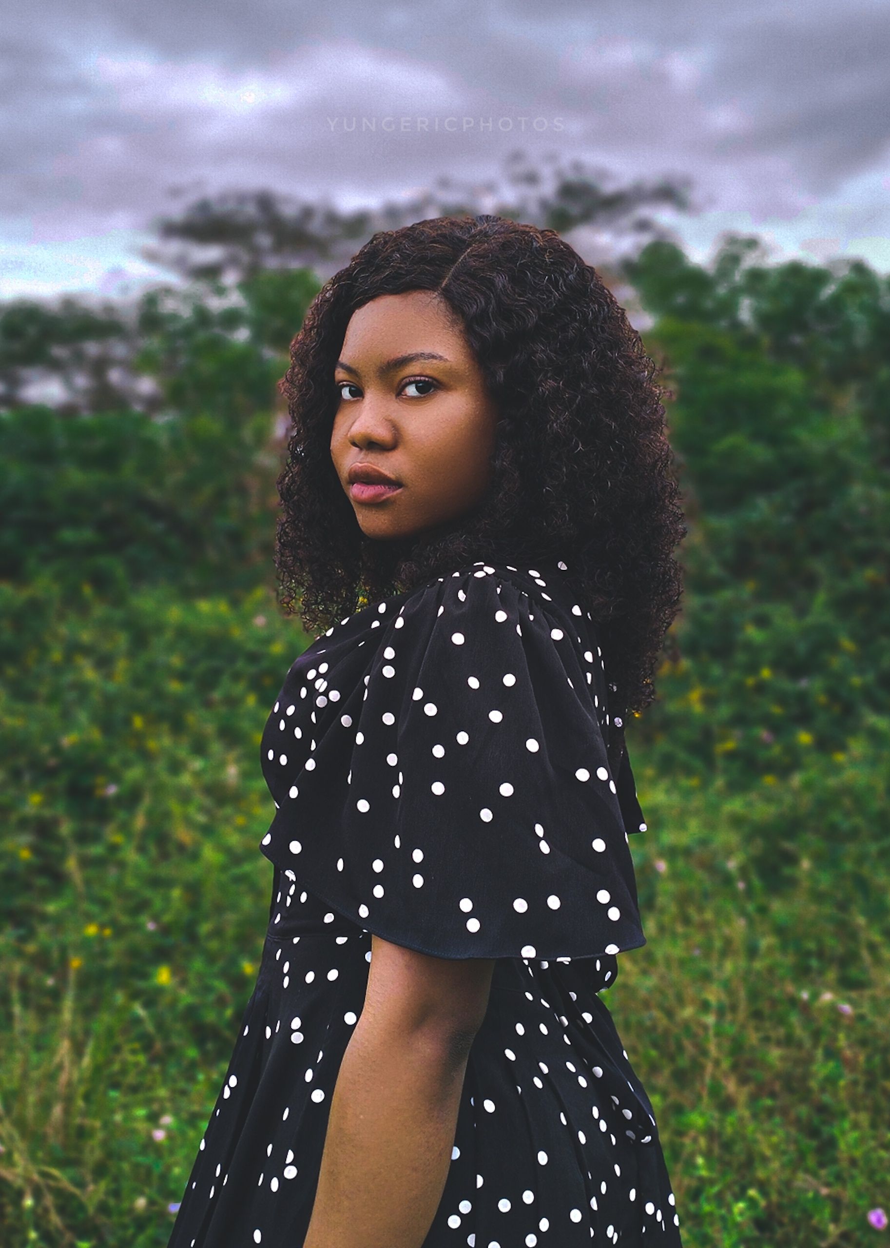 A woman in a black polka dot dress poses in a lush outdoor setting, exuding confidence and style.