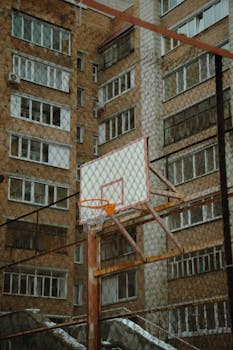 Outdoor basketball hoop amidst urban apartment buildings, fenced court.