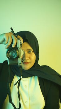 A woman wearing a hijab holding a camera, capturing a moment indoors with a DSLR.