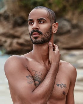 Close-up of a bearded man with tattoos standing shirtless on a beach.