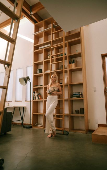 Woman in white pants standing by a tall bookshelf in a stylish, modern library interior.