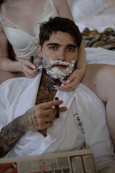 Tattooed man being shaved while holding a cigar, wearing a robe indoors, creating a relaxed atmosphere.