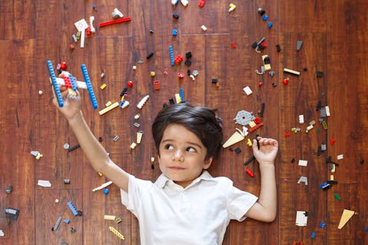 Top view of concentrated kid lying on floor among various details of construction set while playing with handmade airplane