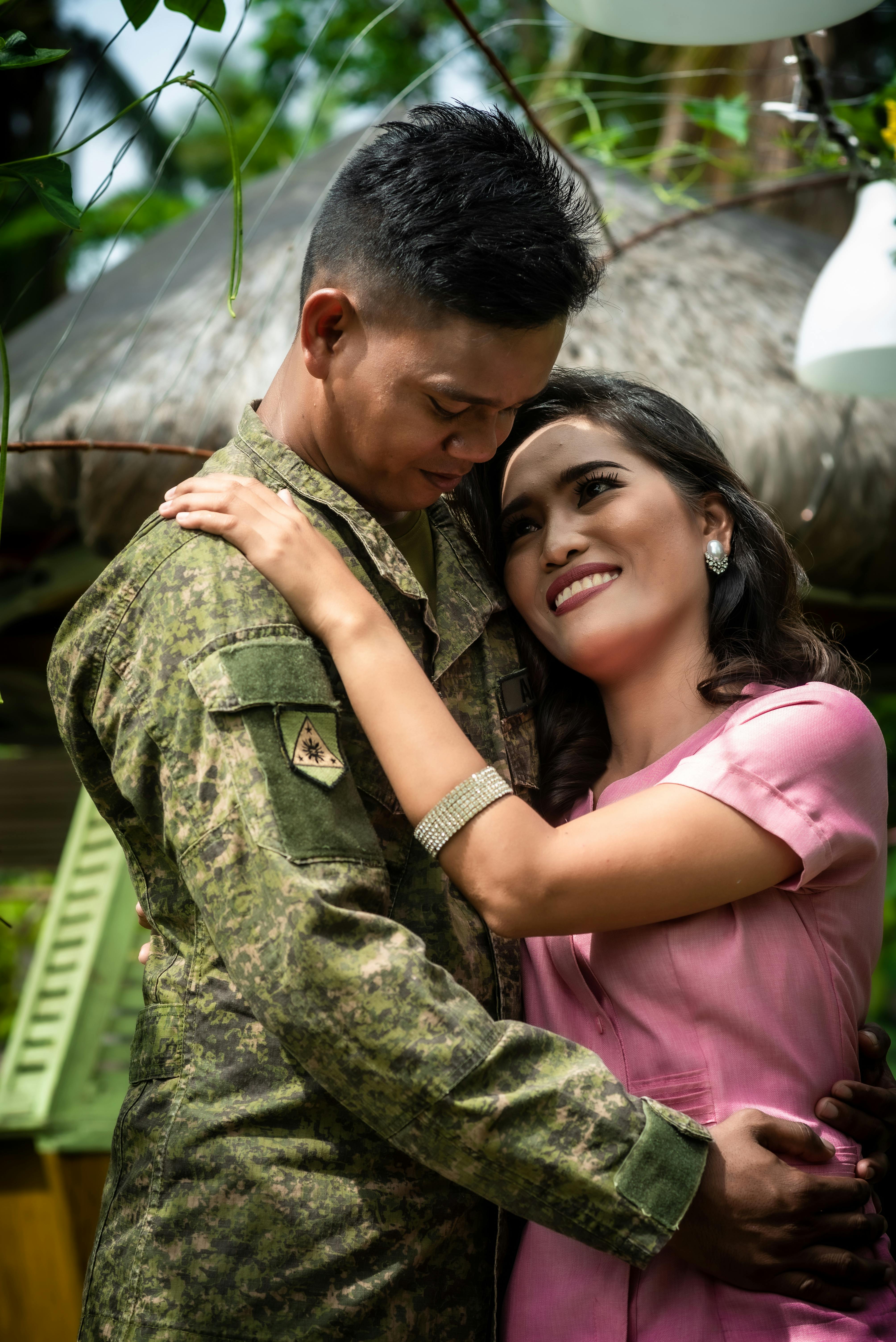 Soldier in camouflage uniform embraces a smiling woman in natural setting.