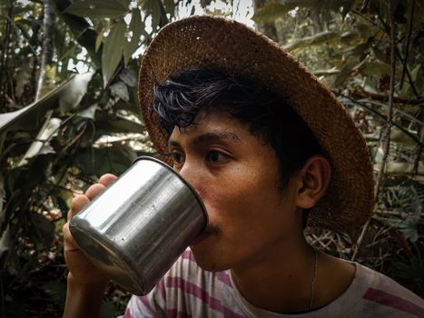 Portrait of an Asian man wearing a straw hat, drinking from a stainless steel cup in the jungle of West Kalimantan.
