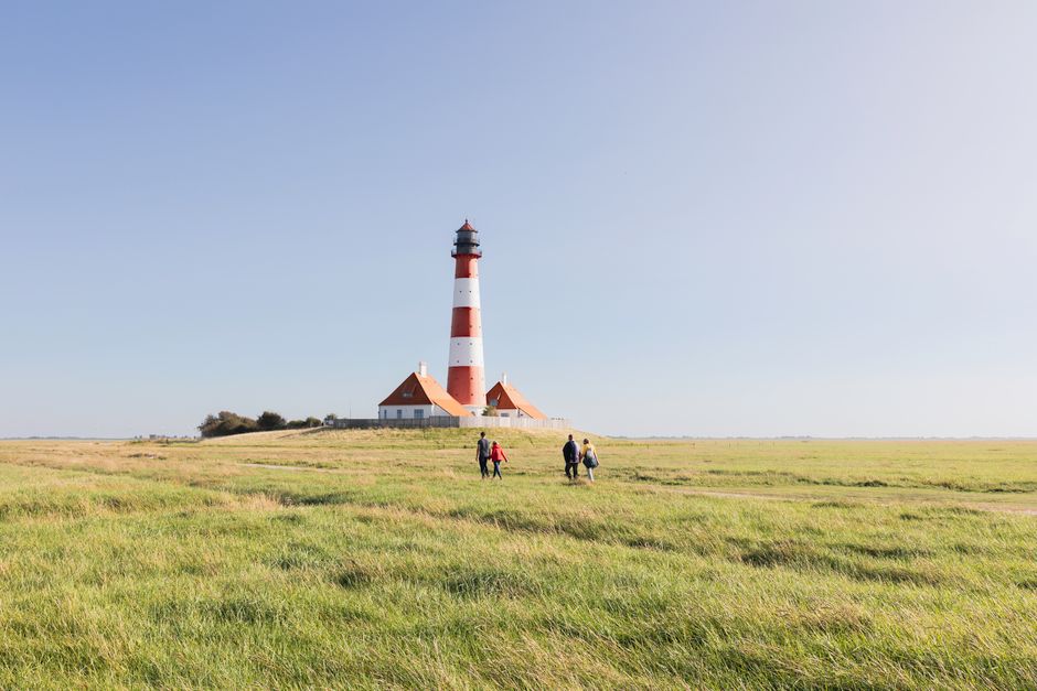 Scenic view of Westerhever Lighthouse with people walking through grassy fields under a clear sky.