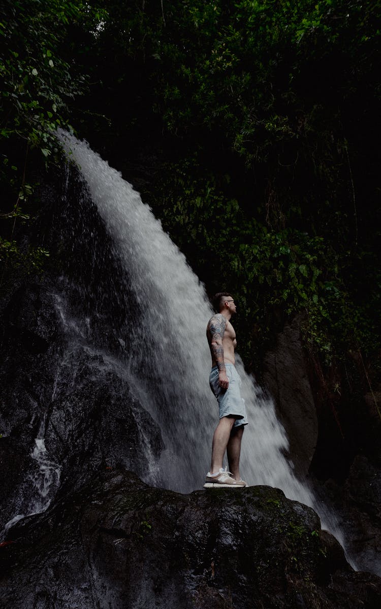 Young Man In Shorts Standing By Waterfall