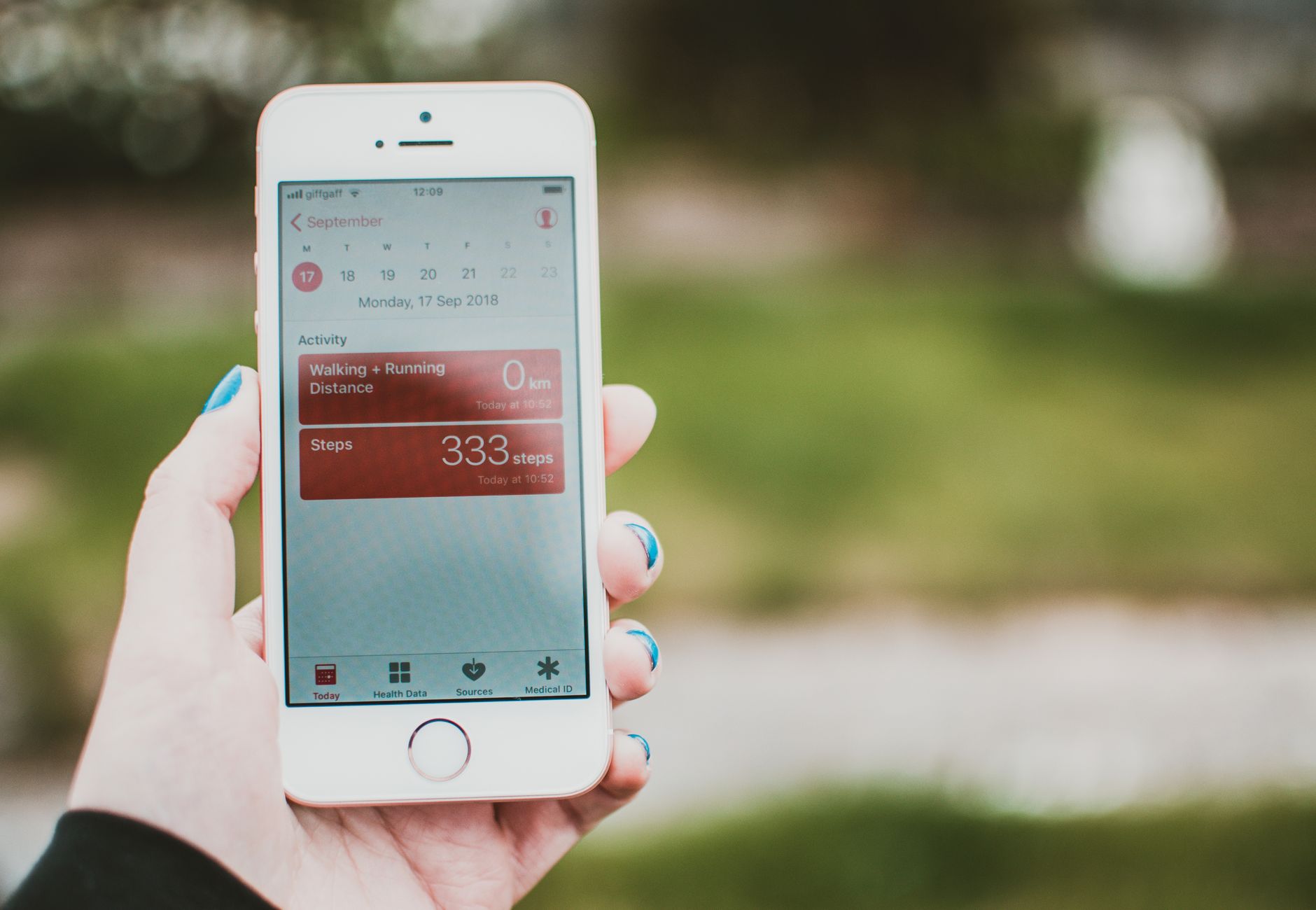 A person holds a smartphone showing a fitness app with step count and distance tracking outdoors.