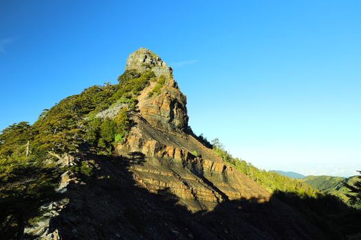 Landscape of a rocky mountain peak with trees under a clear blue sky and bright sunlight.