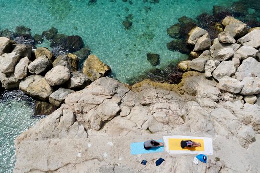 Two women doing yoga on mats by the rocky seaside with clear blue water.