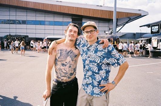 Two men standing together outdoors, one with eyeglasses and tattoos, smiling at a festival venue.