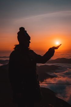 Silhouette of a person holding the sun during sunset on a mountain top, creating a serene and beautiful scene.