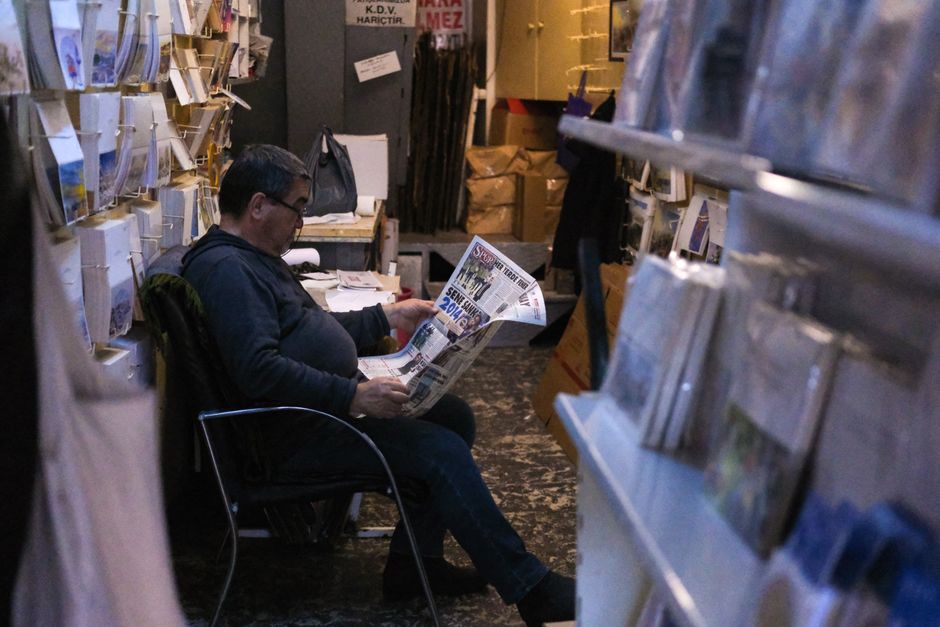 Gentleman sits comfortably reading a newspaper in a cozy, dimly lit bookstore.