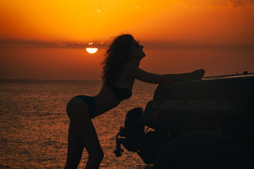 Silhouette of a Woman in a Bikini Leaning against a Car on a Shore at Sunset 