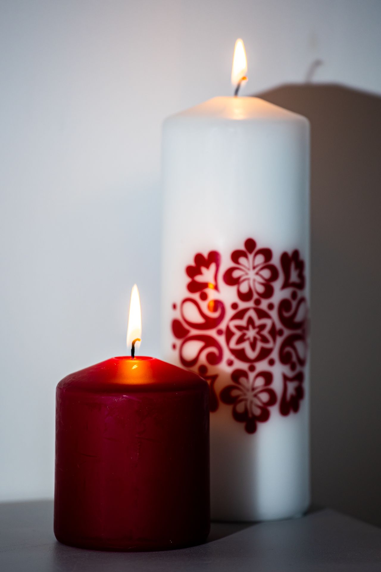 A lit red candle in front of a tall white candle with a red floral pattern